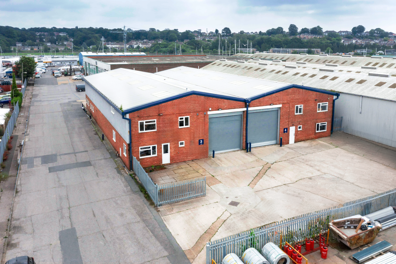 Aerial view of a red-brick industrial warehouse with two large grey roller shutters and adjacent car park, surrounded by a metal fence.