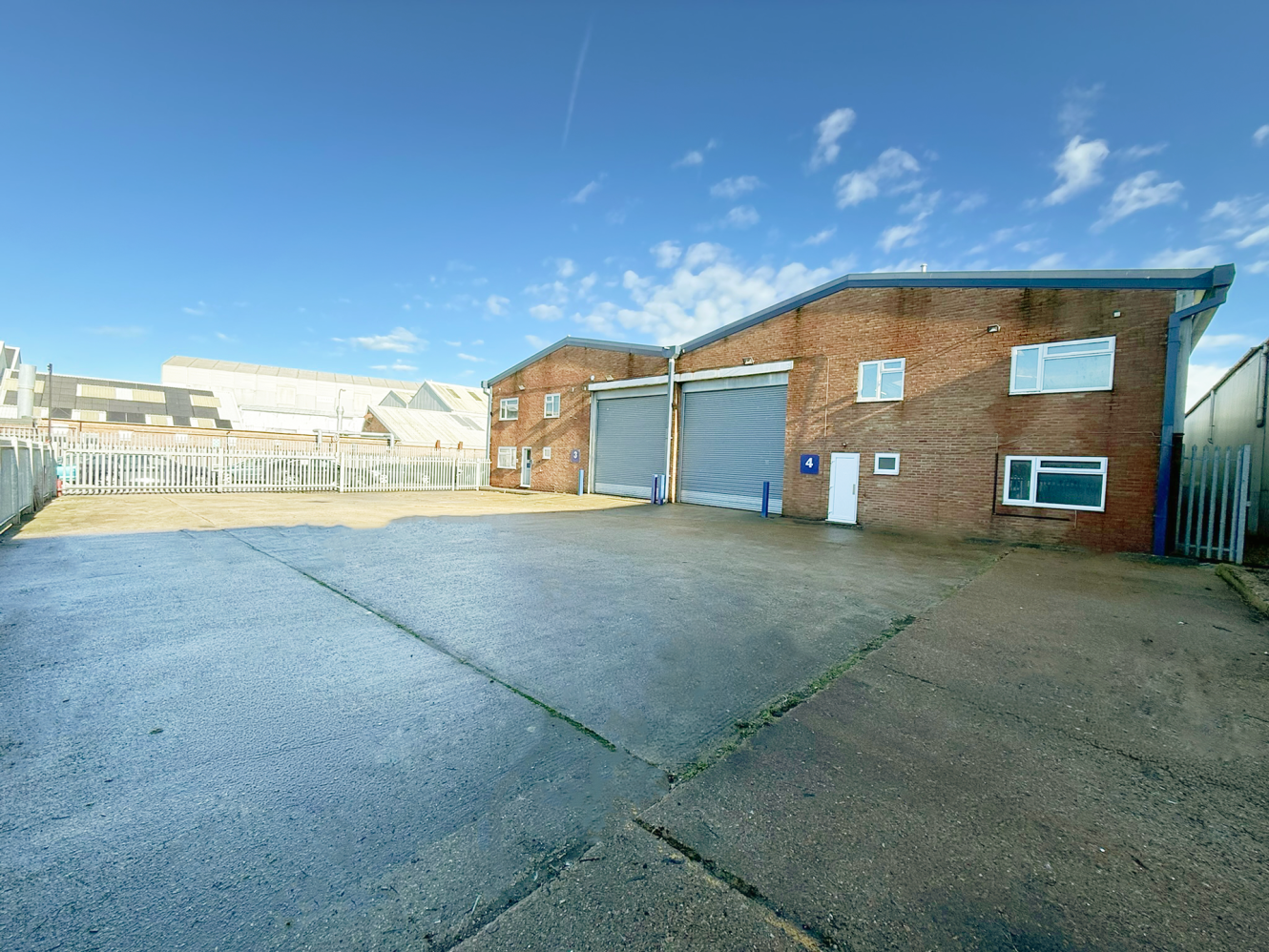 A large commercial or industrial building with two roller shutter doors, brick exterior, and a wide, empty paved forecourt under a clear sky.