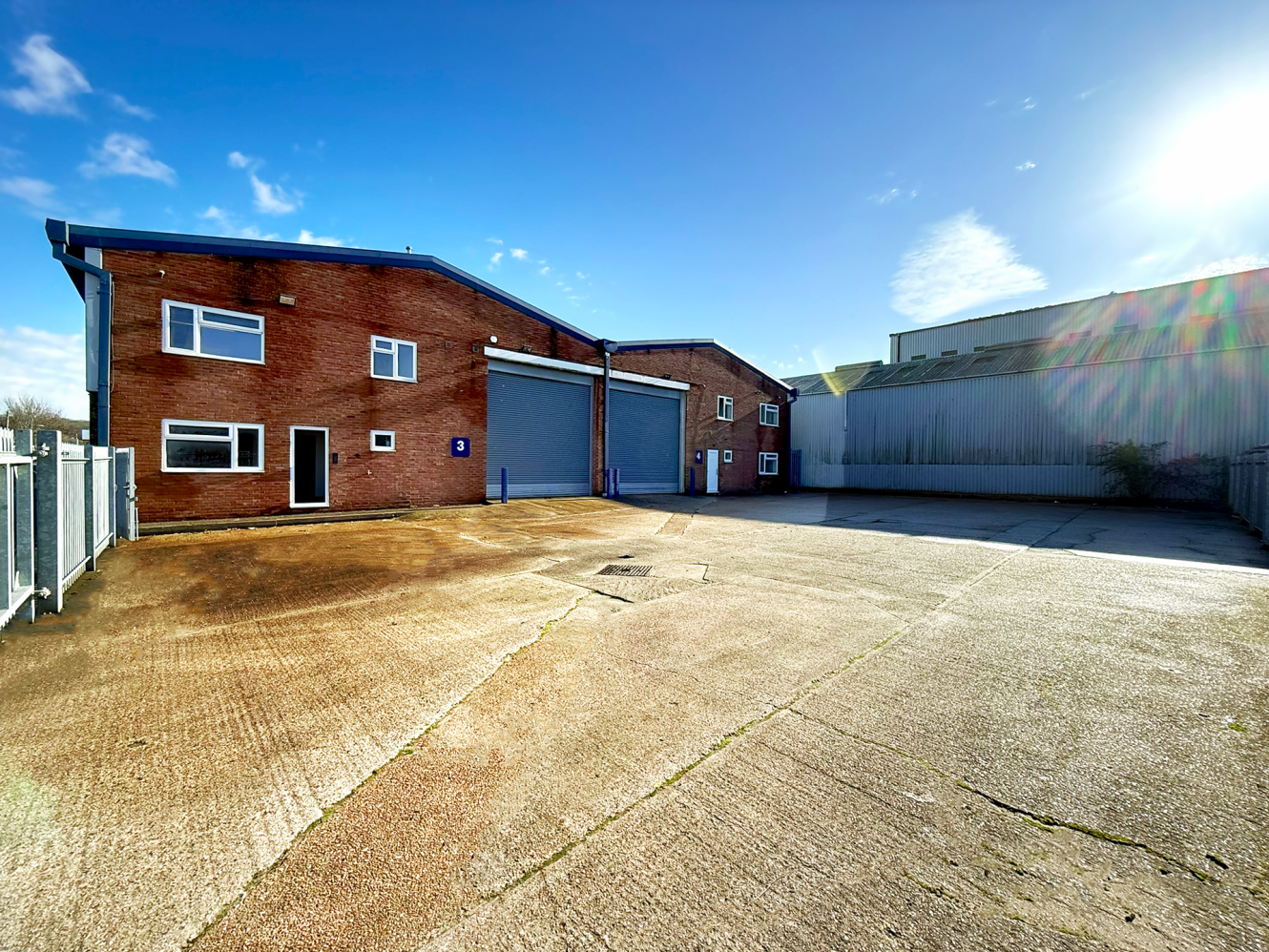 A brick industrial building with two roller shutter doors, windows, and a fenced concrete yard under a clear blue sky with sunlight.