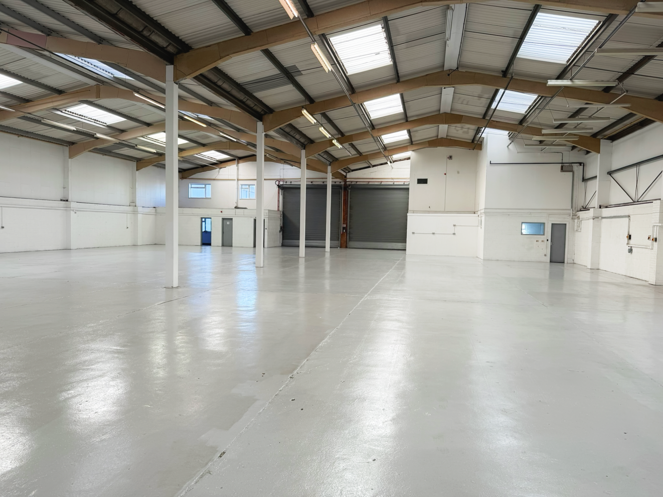 Spacious, empty industrial warehouse with white walls, high ceilings, exposed beams, and large roller shutter doors. Polished concrete floor and several windows provide natural light.