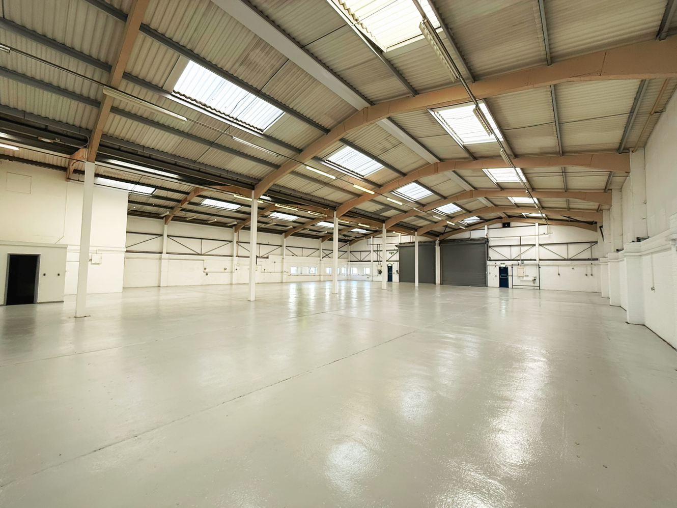 Large, empty warehouse with a polished concrete floor, high ceilings, exposed beams, skylights, and several doors along white walls.