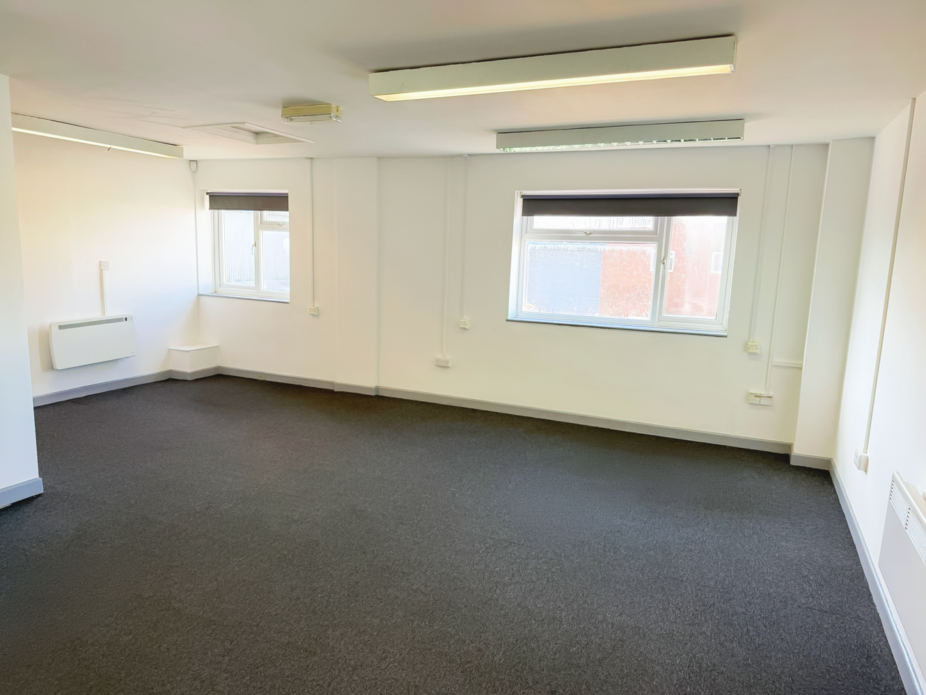Empty office room with white walls, two windows with black blinds, grey carpet flooring, and ceiling lights.