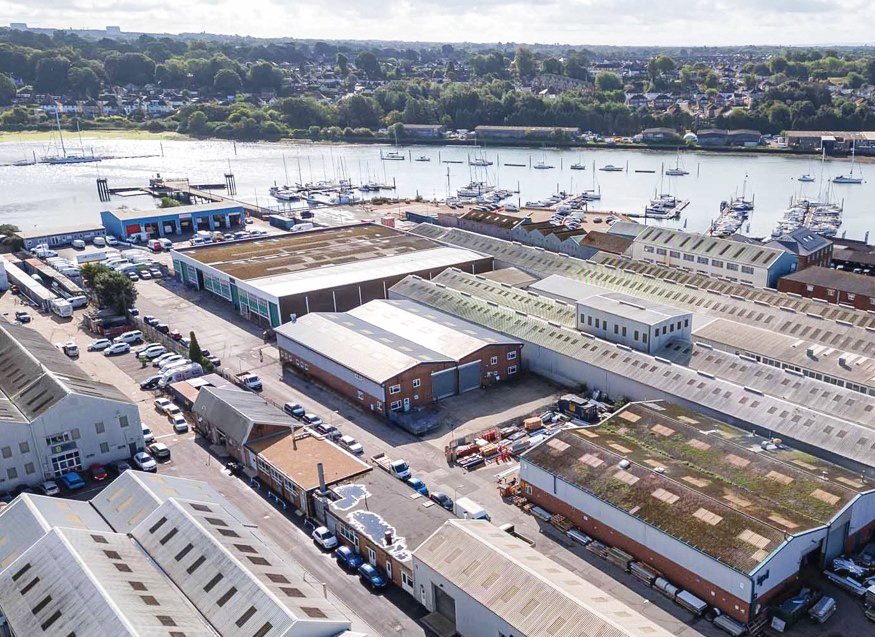 Aerial view of an industrial area with warehouses and parked cars near a river with moored boats and a residential neighbourhood in the background.