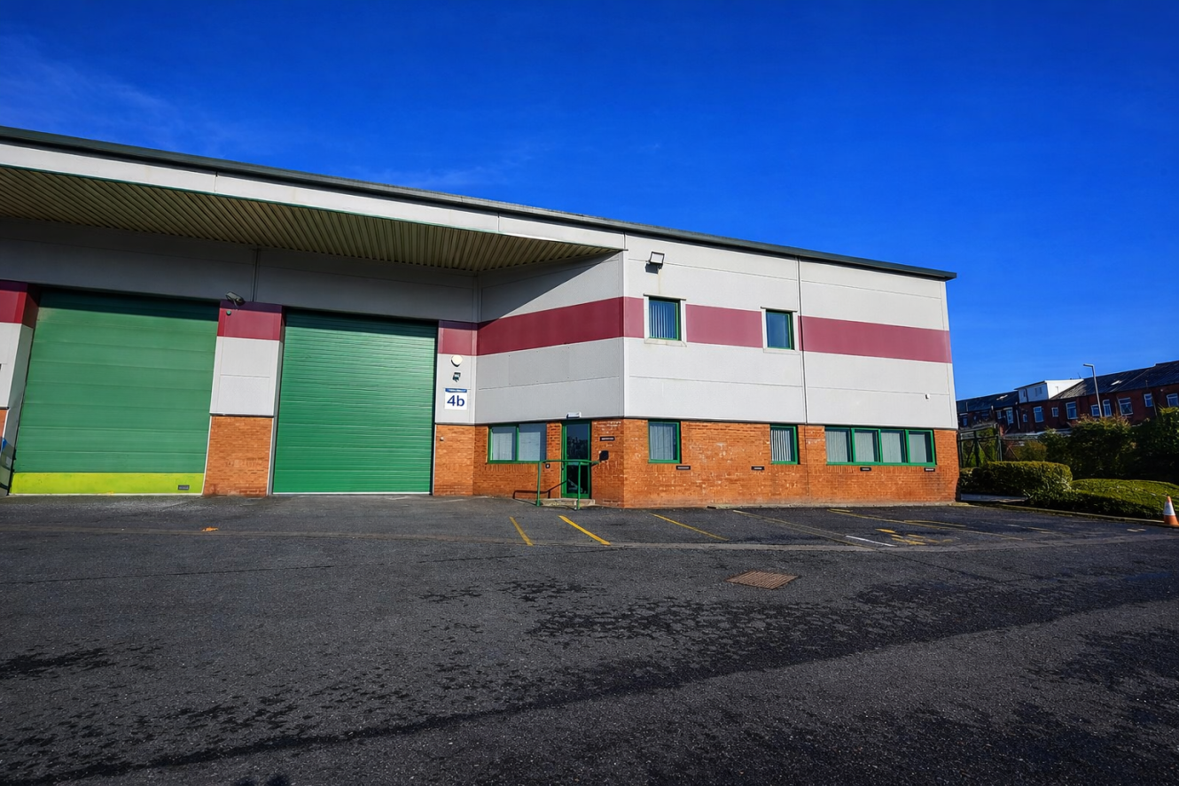 Industrial warehouse building with large green doors, white and red-striped upper façade, brick lower section, and empty parking spaces in front under a clear blue sky.