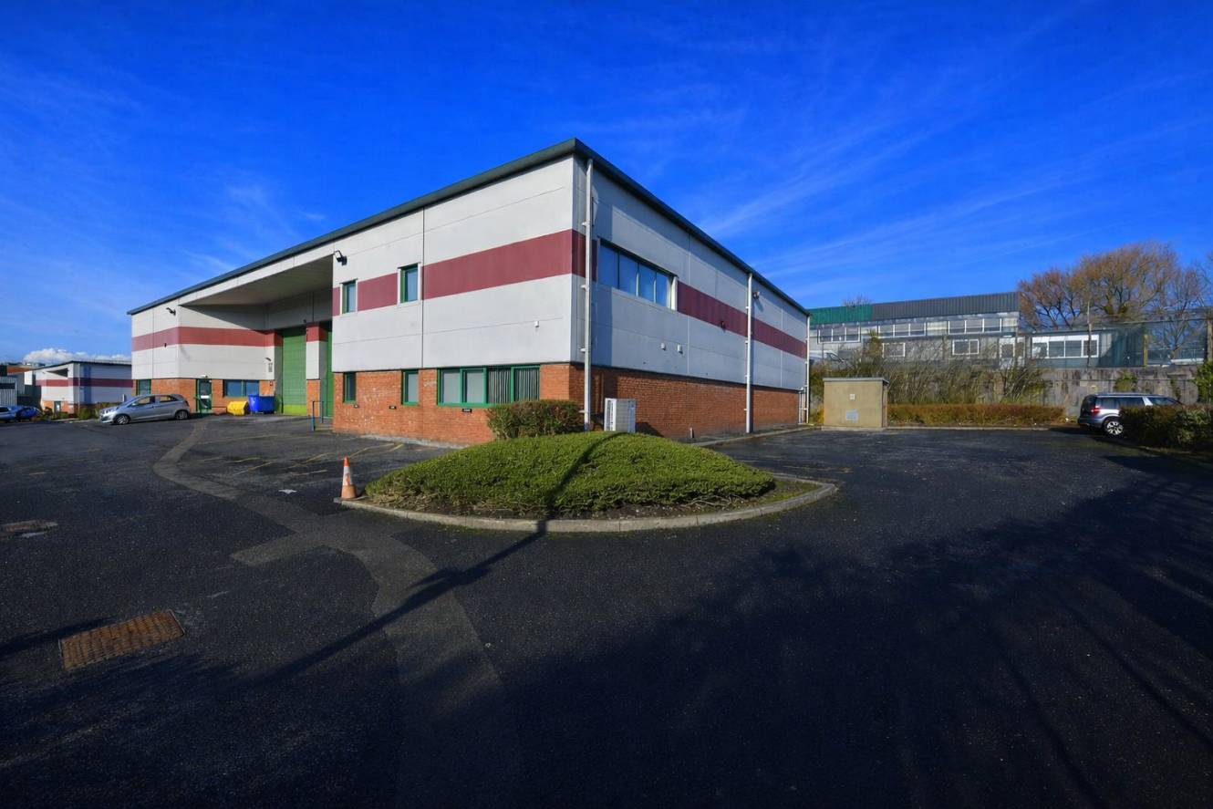 A large industrial building with red and grey stripes, surrounded by a paved car park and sparse greenery under a clear blue sky.