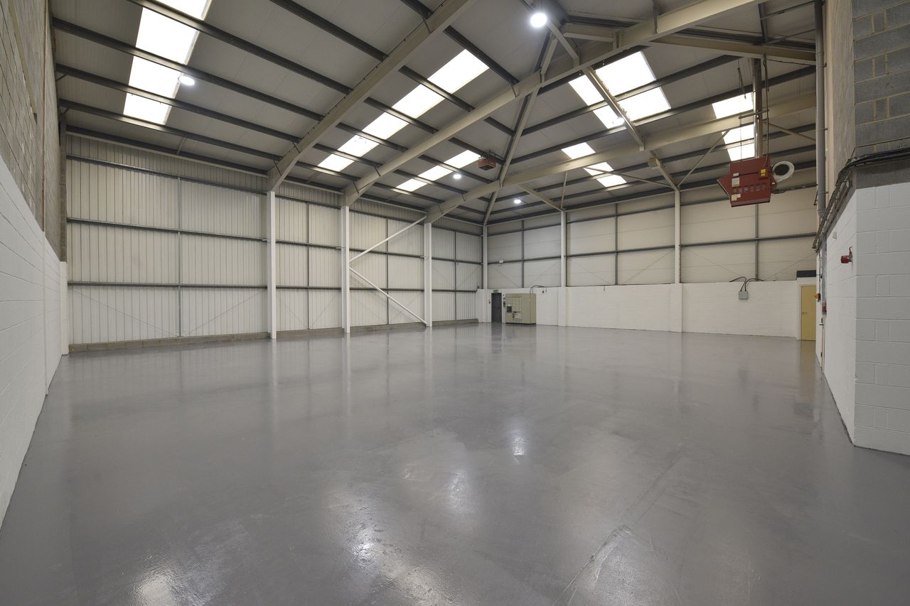 Empty industrial warehouse with high ceilings, exposed beams, white walls, grey floor, and fluorescent lighting.