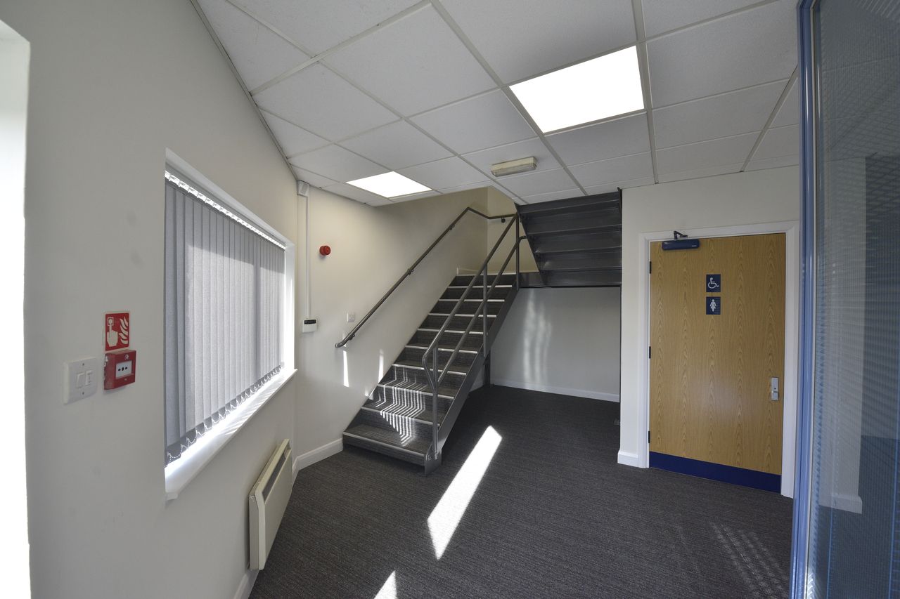 A modern indoor stairwell with metal steps, grey carpet, a window with blinds, a fire alarm, and a closed door with a toilet sign. Ceiling lights illuminate the space.