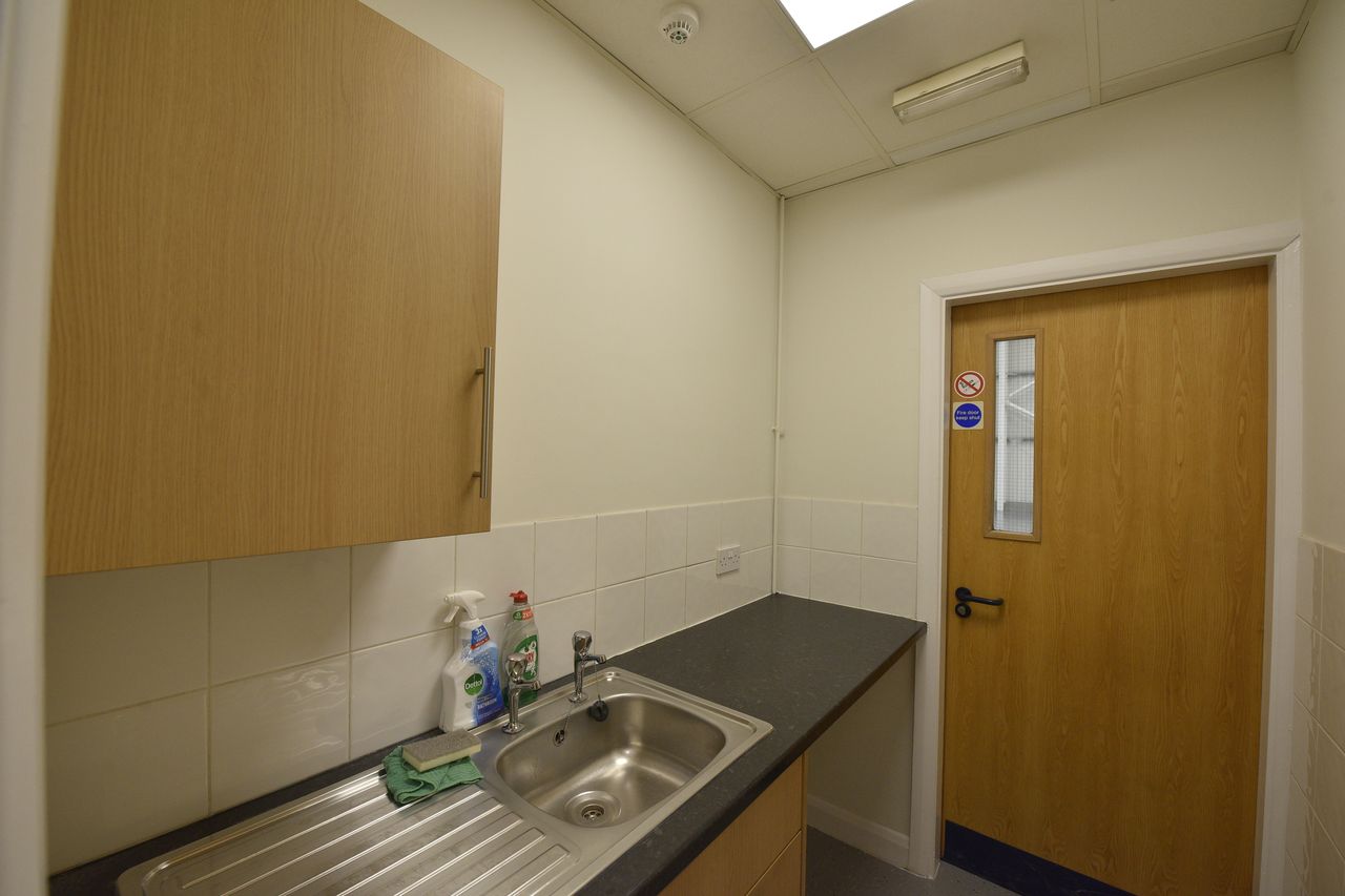 A small kitchenette with a stainless steel sink, worktop, cleaning supplies, wooden cupboards, tiled splashback, and a closed wooden door with a window.