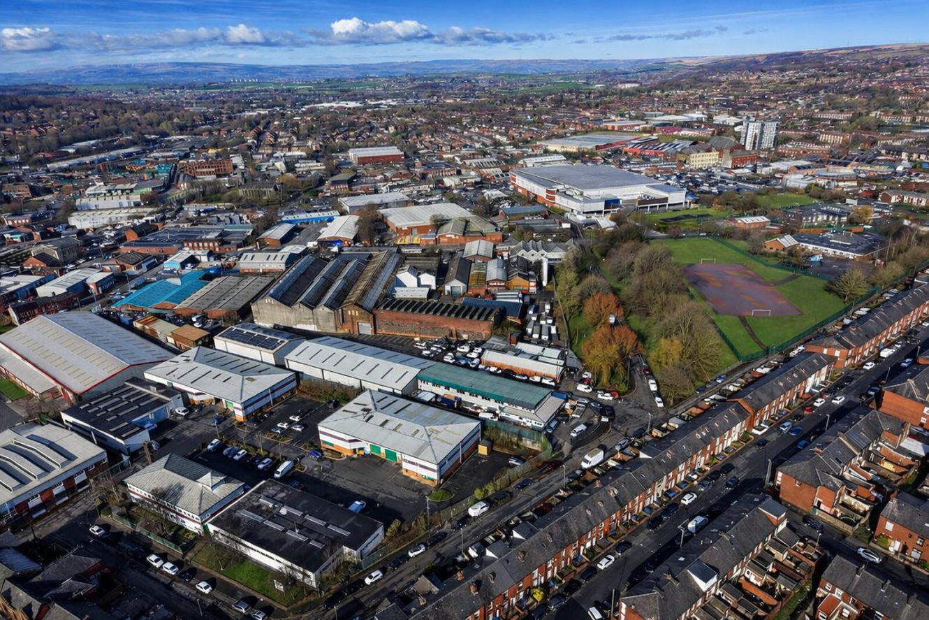Aerial view of an urban area with warehouses, industrial buildings, residential houses, tree-lined streets, and a large sports pitch. Hills and countryside are visible in the background.