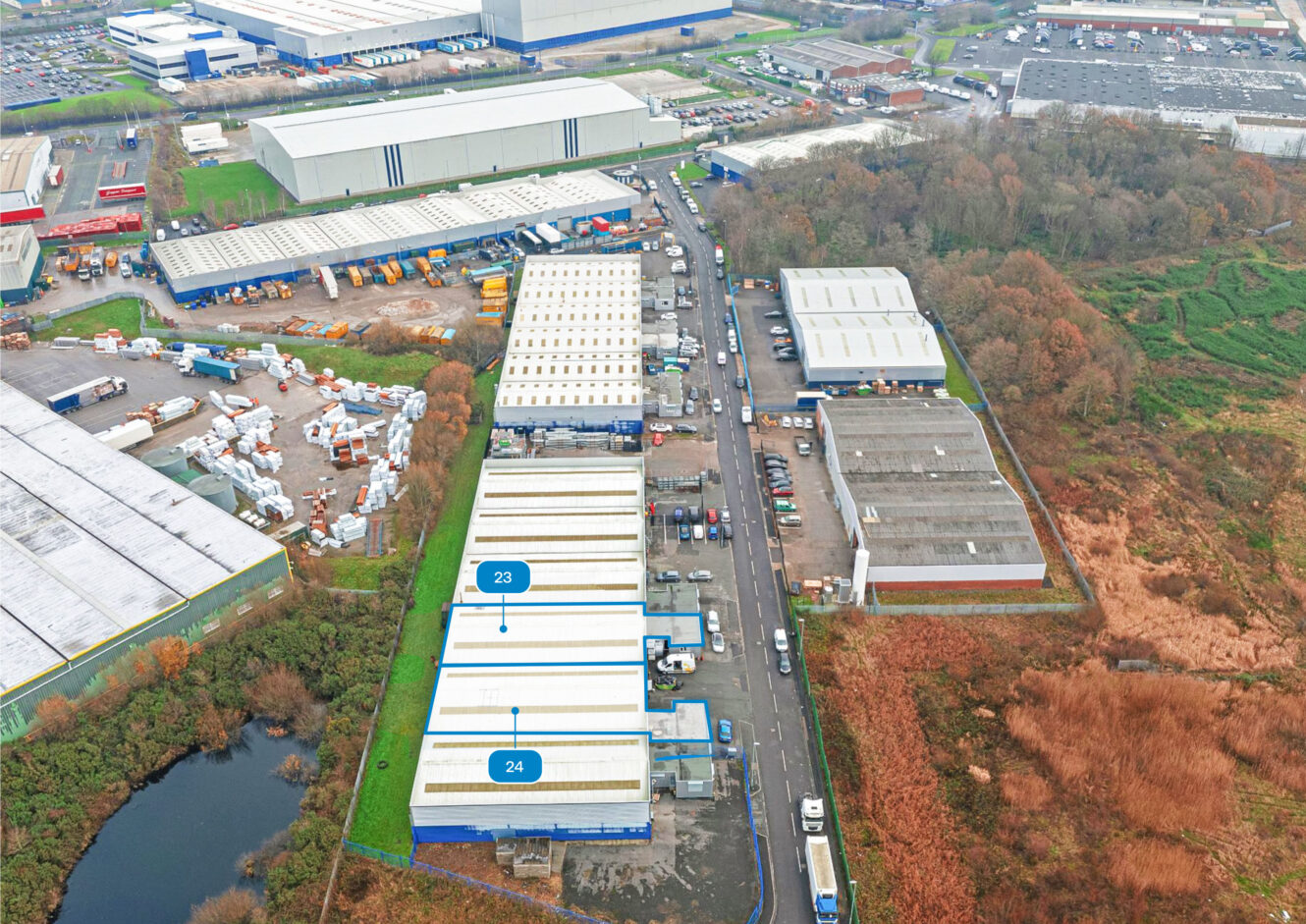 Aerial view of an industrial estate with warehouses, labelled units 23 and 24, adjacent to a road with parked vehicles and surrounding greenery.