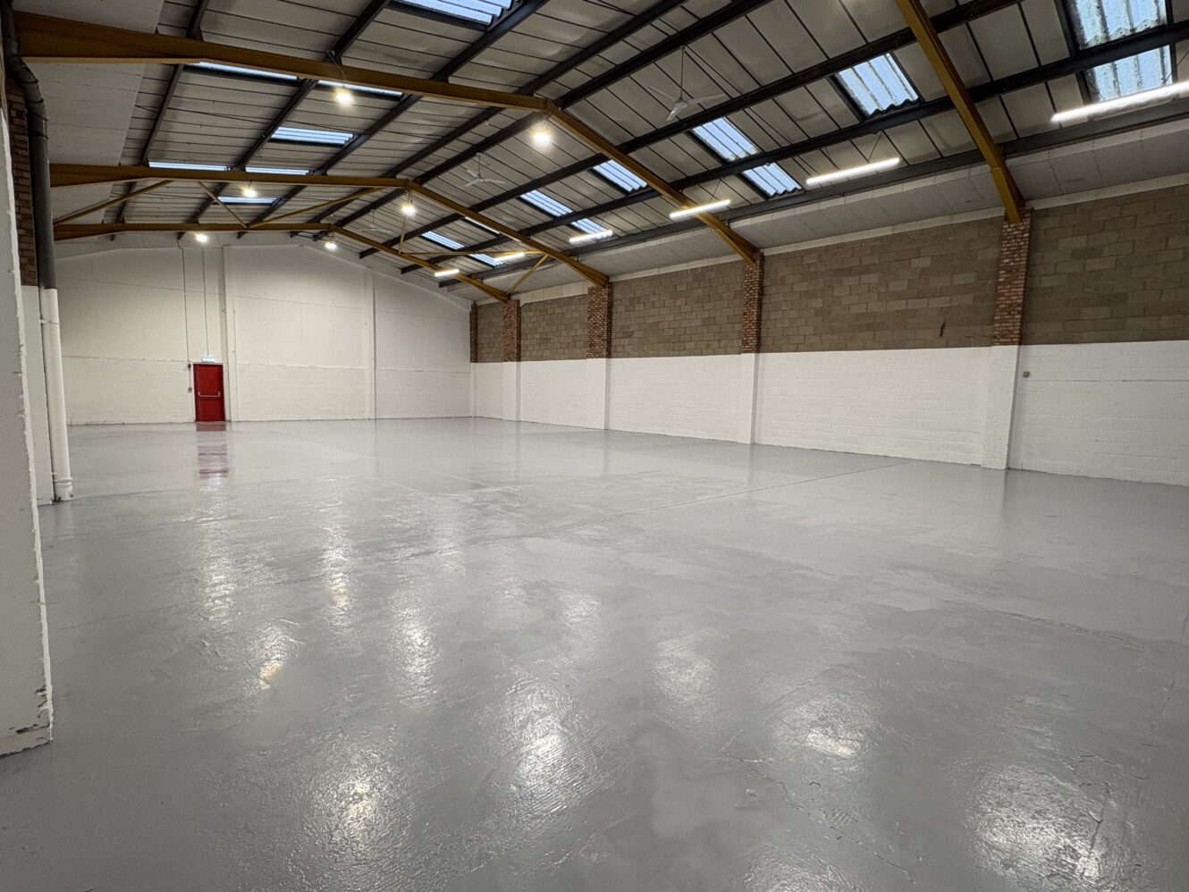 Interior of an empty industrial warehouse with a grey concrete floor, high ceiling, exposed beams, brick and white painted walls, and a red door at the far end.