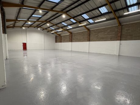 Interior of an empty industrial warehouse with a grey concrete floor, high ceiling, exposed beams, brick and white painted walls, and a red door at the far end.