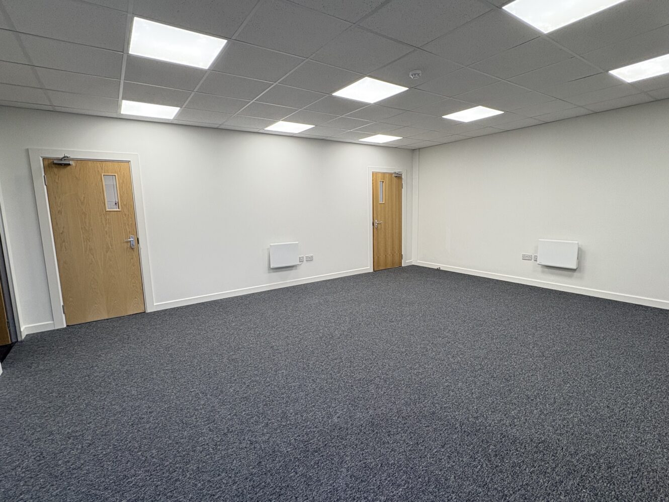 An empty room with white walls, a grey carpeted floor, and two closed wooden doors. The ceiling has recessed lighting panels.