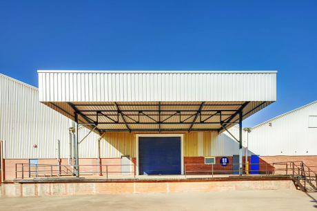 A loading bay with a metal roof, brick lower walls, a blue roller shutter door, and a ramp, under a clear blue sky.