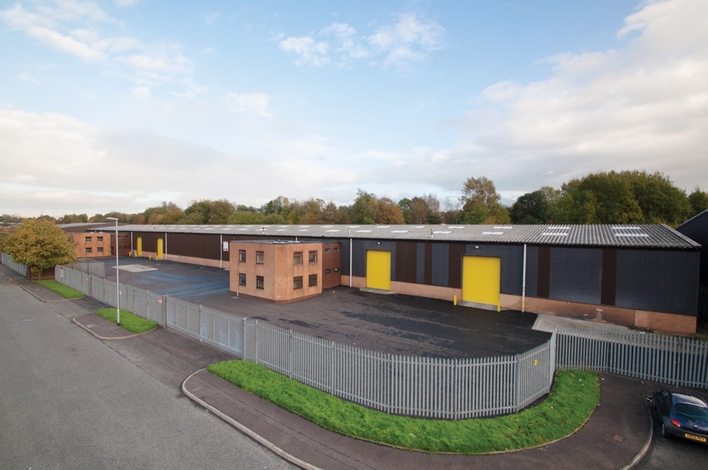 A large industrial warehouse with brown and black exterior walls, yellow doors, and an adjacent office building, surrounded by a metal fence and situated on a quiet street.
