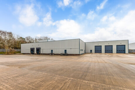 Large industrial warehouse with multiple loading bays and garage doors, set on a wide concrete yard under a partly cloudy sky.