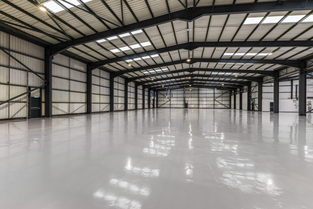 Large empty industrial warehouse with high ceilings, steel beams, polished concrete floor, and overhead lights.