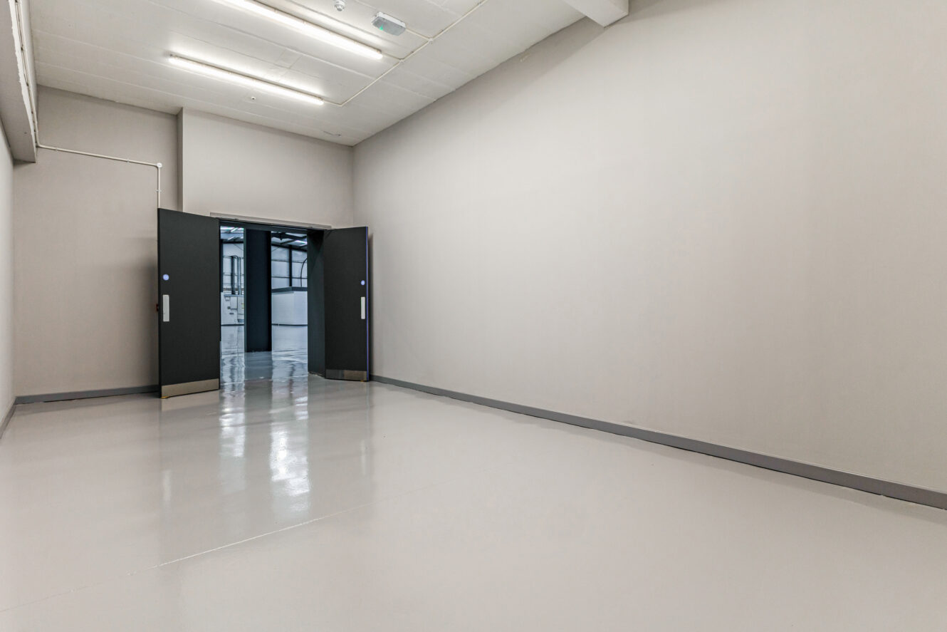 Empty room with smooth light-coloured flooring, plain walls, and double doors at the far end opening to a brighter adjacent space.