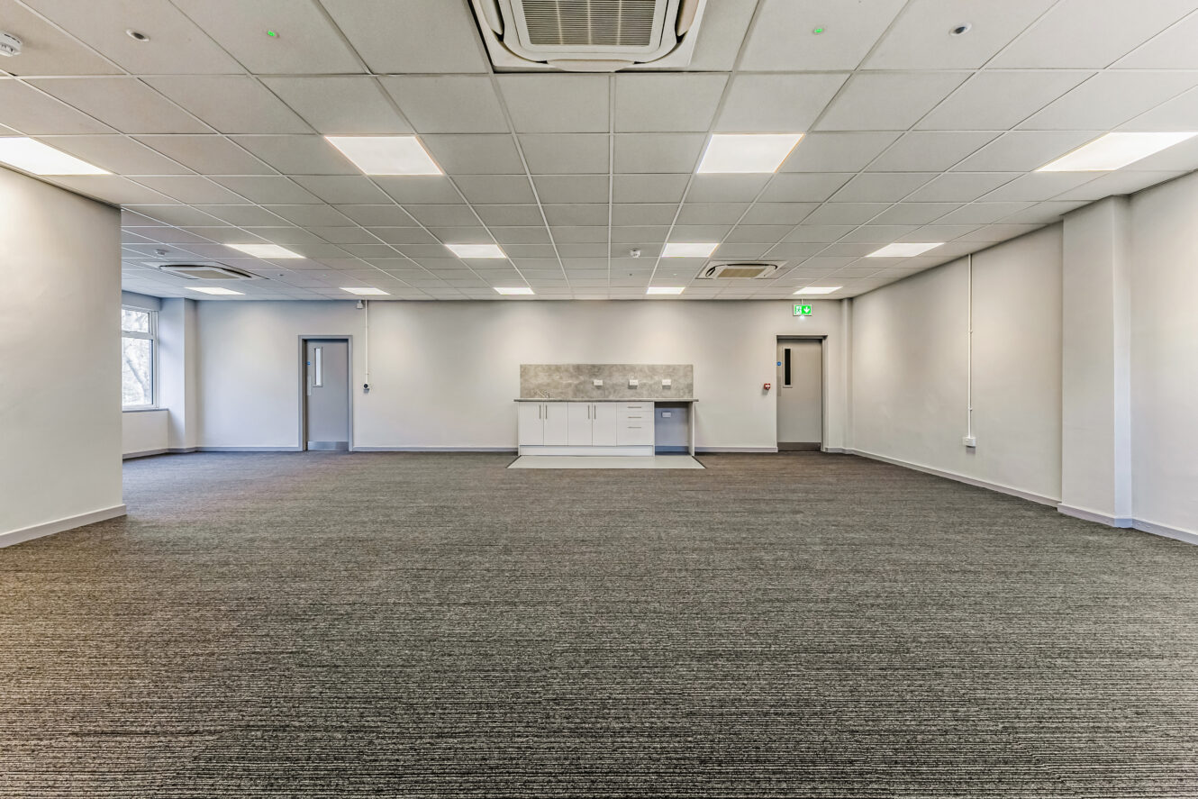 Spacious empty office room with grey carpet, white walls, ceiling lights, a small kitchenette, and two doors, one with a window and one near a green exit sign.