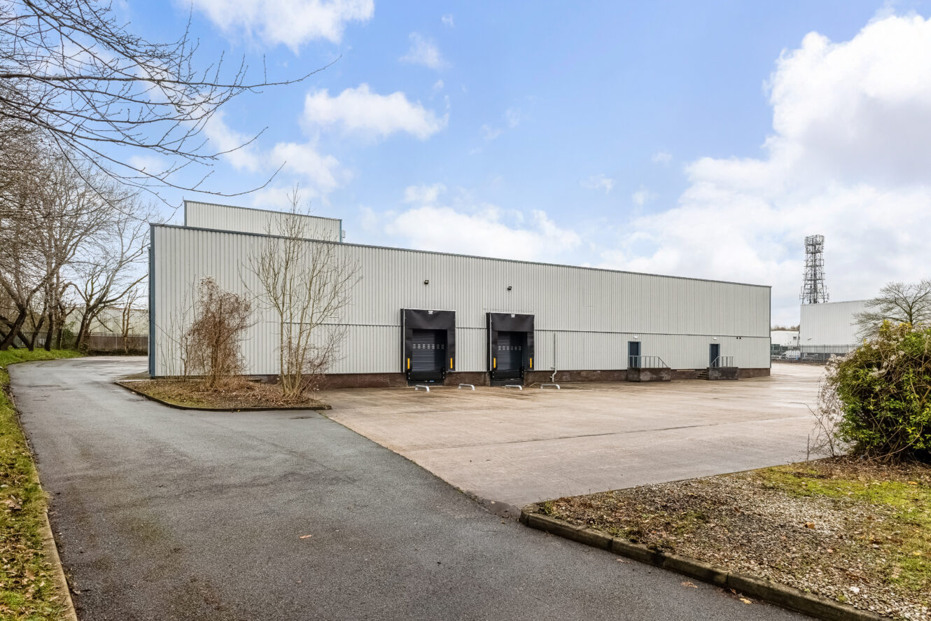 A large white industrial warehouse with two loading bays, surrounded by a tarmacked area and minimal landscaping, under a partly cloudy sky.