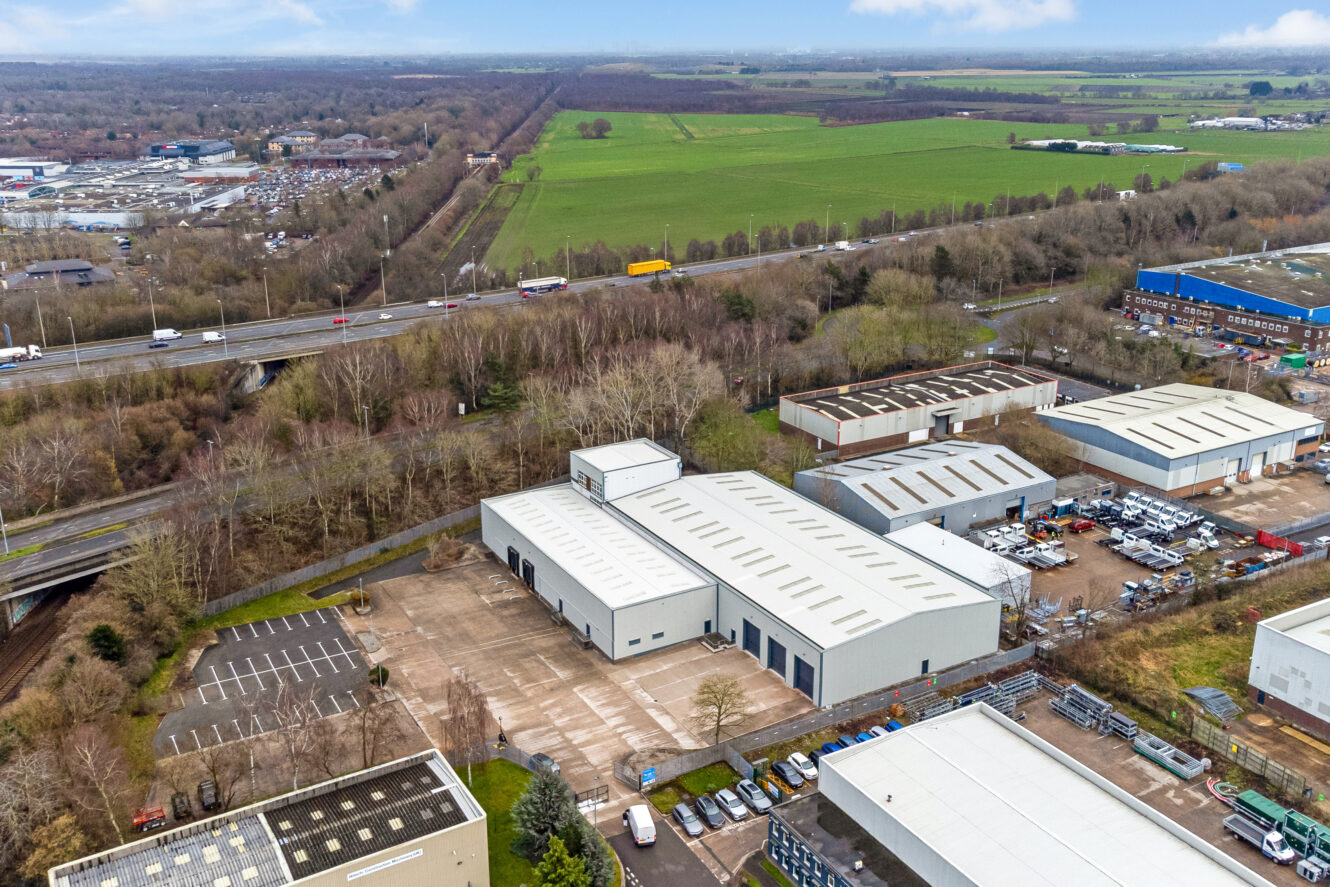 Aerial view of industrial warehouses with car parks, adjacent to a motorway, surrounded by fields and trees.