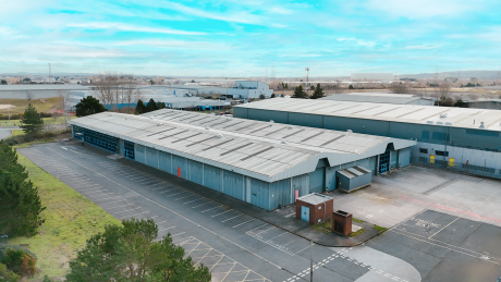 A large industrial warehouse with a grey roof and exterior, bordered by an empty car park and greenery under a clear blue sky.