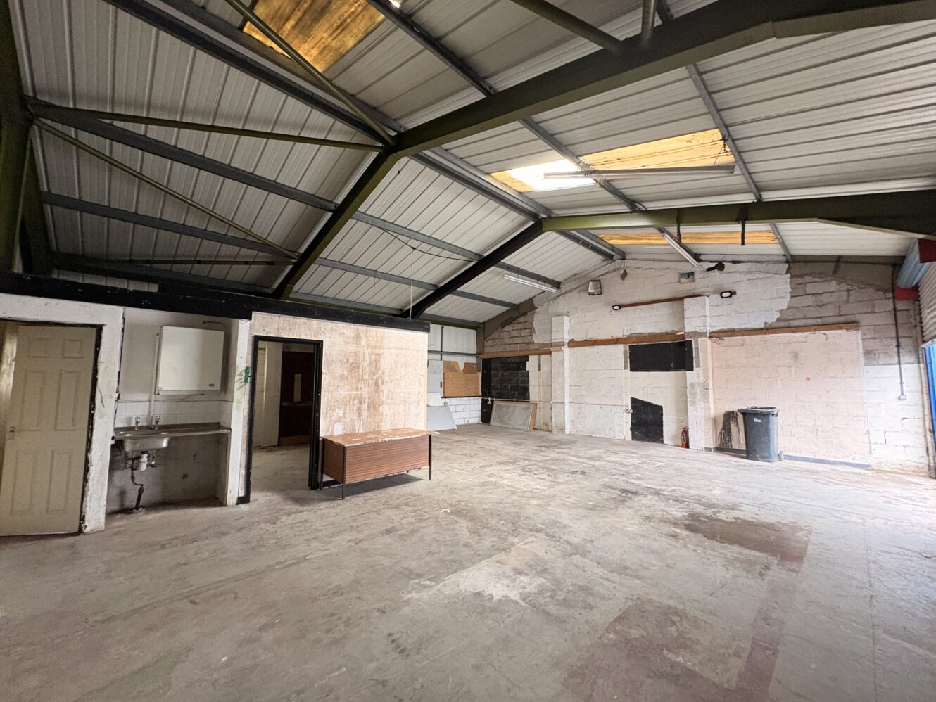 Large, empty industrial room with a high metal ceiling, exposed walls, a bench, rubbish bin, and sink near a door on the left side.