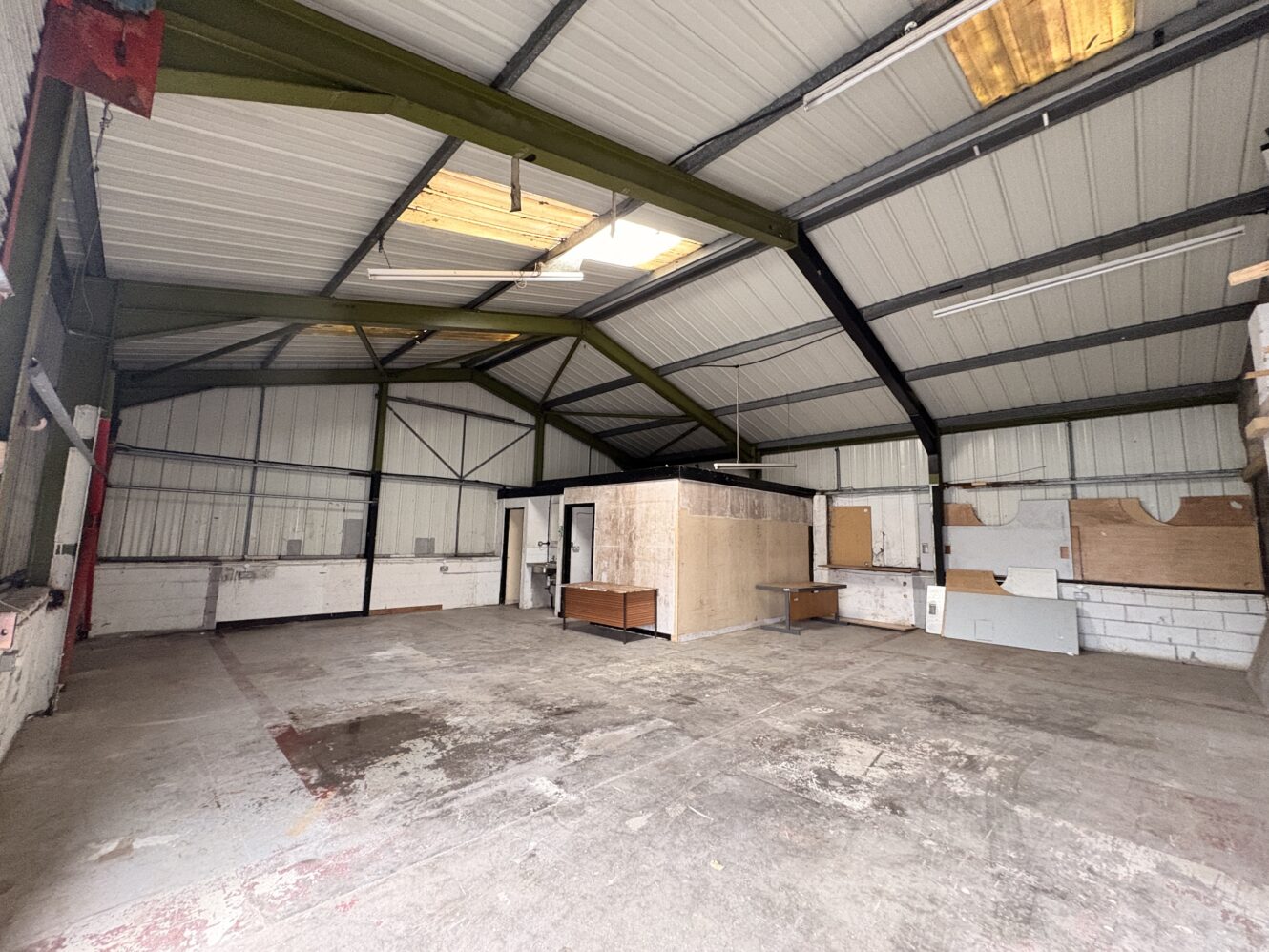Empty warehouse interior featuring exposed metal beams, concrete floor, a small office partition, and scattered rubbish.