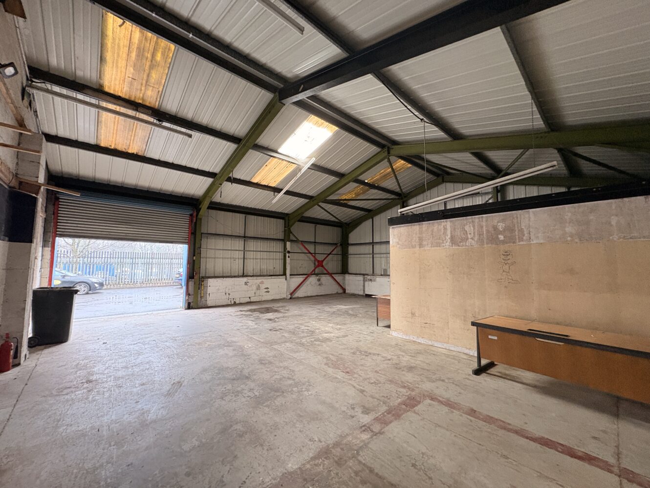 Empty industrial warehouse with a high metal ceiling, open roller shutter door, concrete floor, wooden partition, and a single desk by the wall.