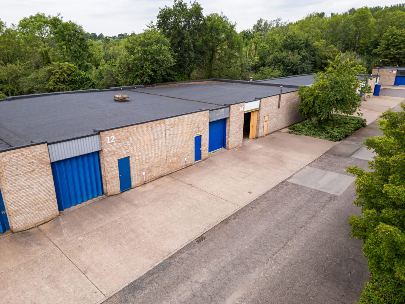 A row of single-storey industrial warehouse units with blue doors and brick walls, trees nearby, and a paved drive in front.