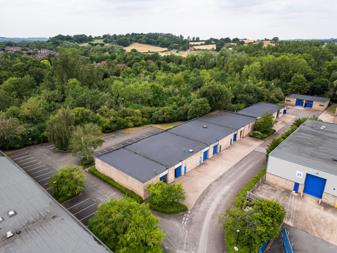 Aerial view of small industrial units with blue doors, surrounded by trees and greenery, set against a rural landscape.