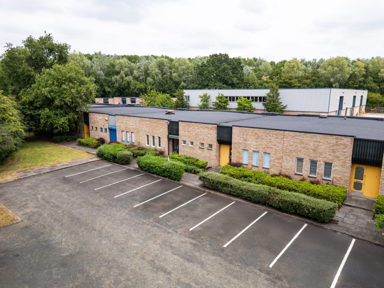 A row of modern, single-storey commercial buildings with yellow doors, surrounded by greenery and an empty car park beneath a cloudy sky.