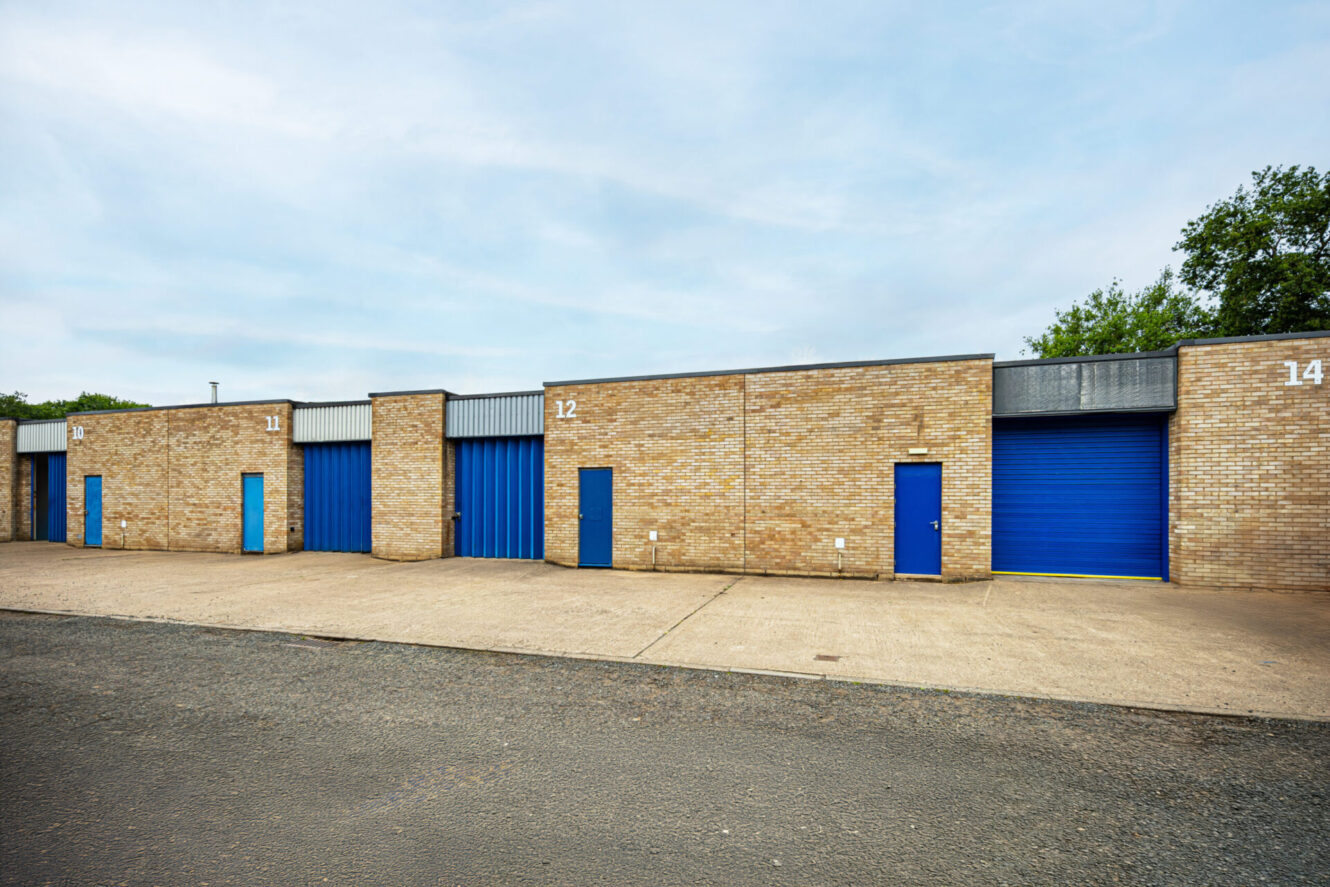 Row of modern industrial storage units with brick walls and blue doors, each unit numbered, on a paved car park under a cloudy sky.