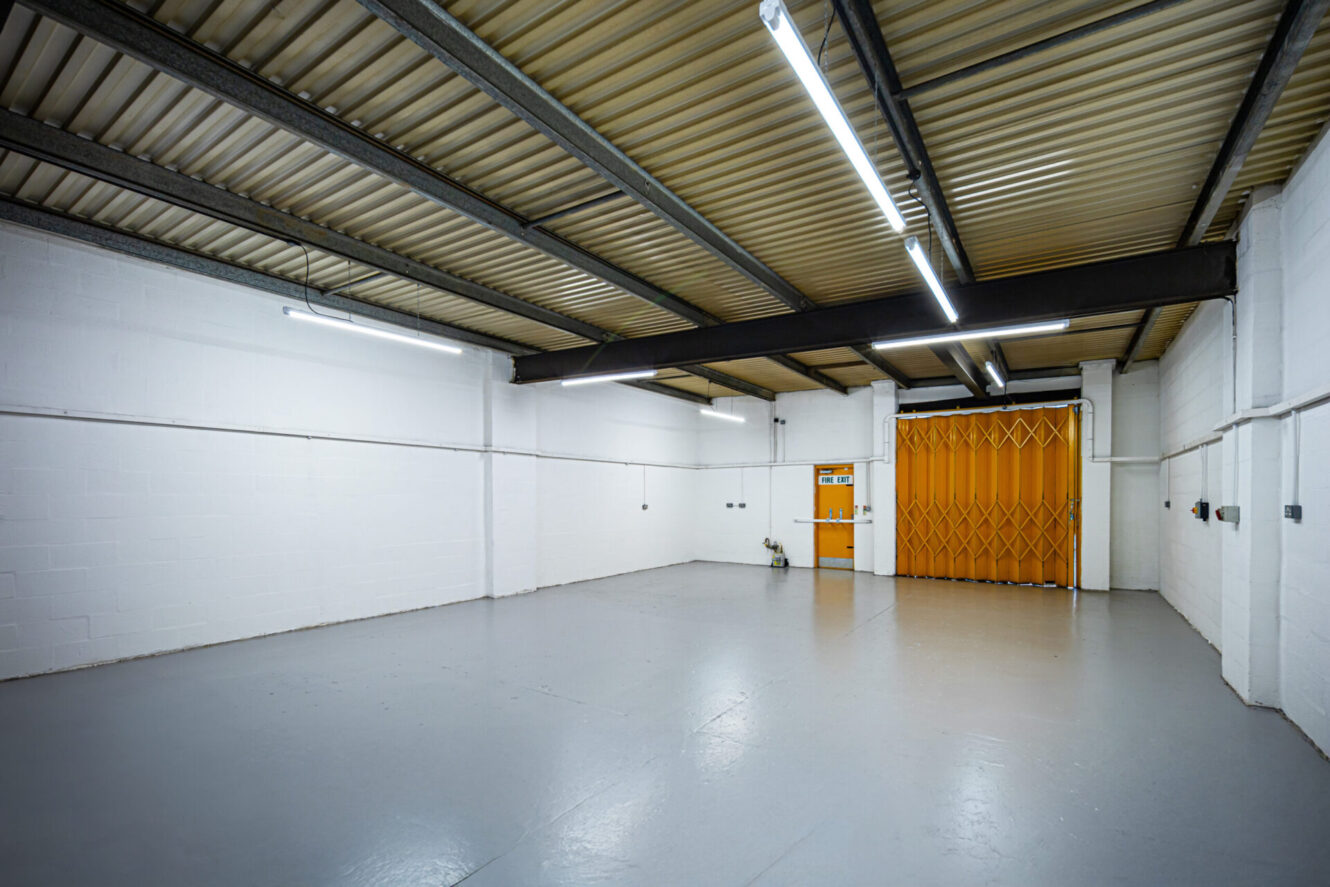 Empty industrial warehouse with white walls, grey concrete floor, fluorescent lights, and a yellow sliding gate at the far end.