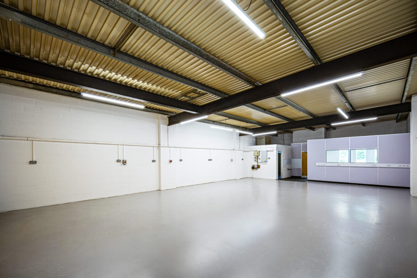 Large, empty industrial room with white walls, exposed metal ceiling beams, fluorescent lighting, and windows above cupboards.