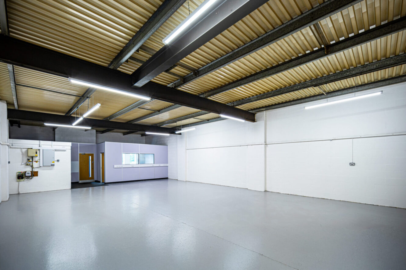 Spacious industrial unit featuring white brick walls, grey epoxy floor, exposed beams, fluorescent lights, and office area at the back.