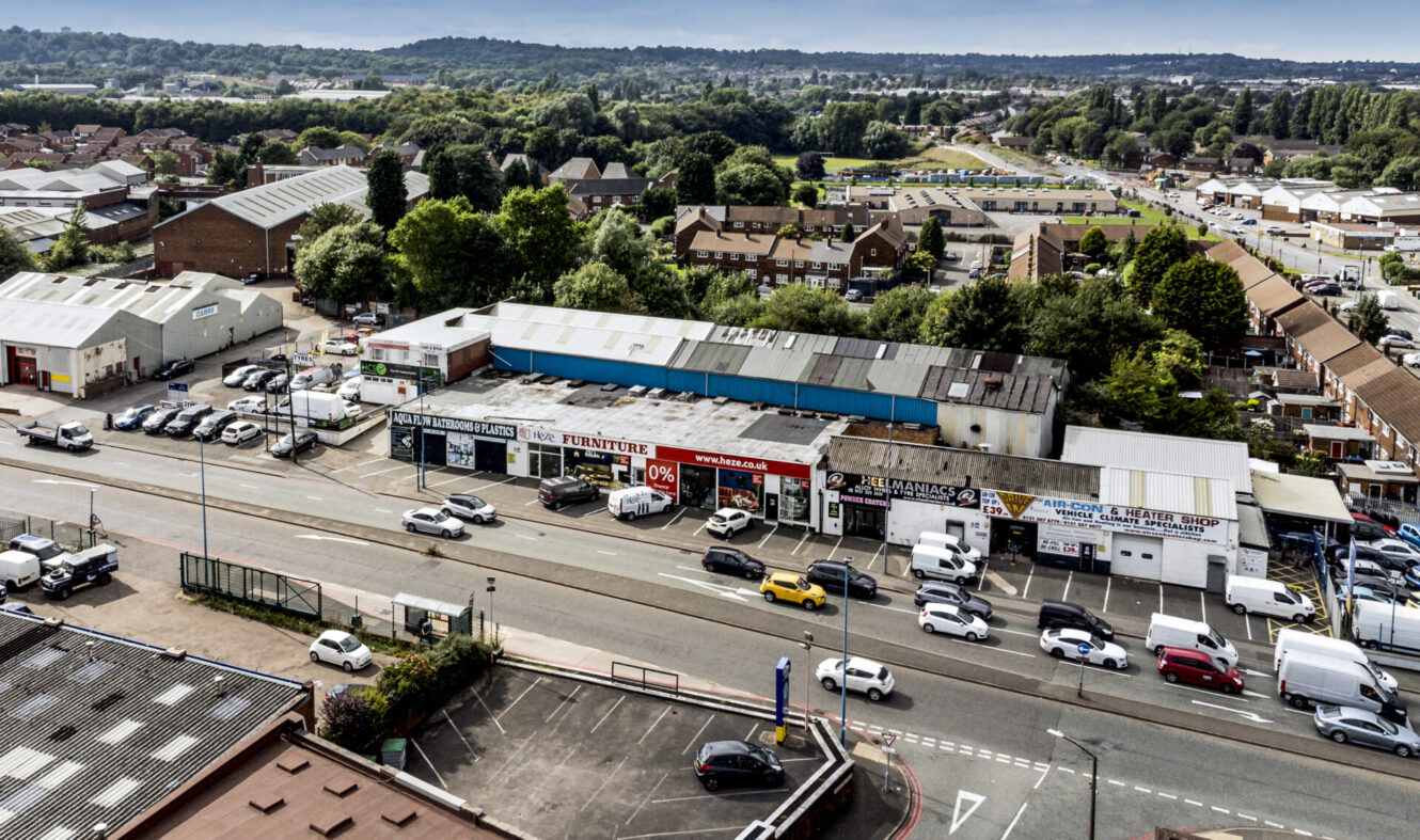 Aerial view of a commercial street lined with shops, cars on the road and parked, and residential buildings with greenery nearby.