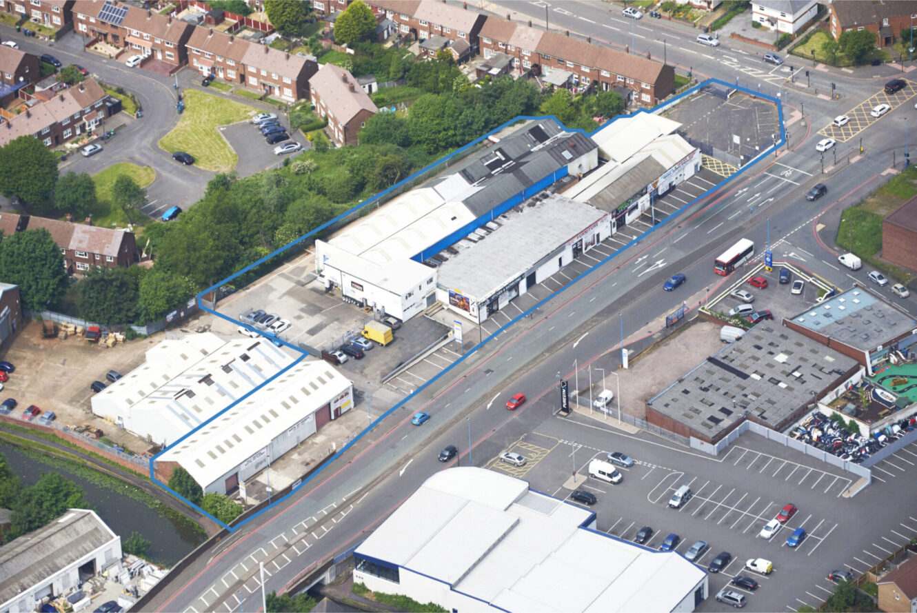 Aerial view of an area featuring warehouses, car parks, a canal, and homes, all split by a central main road.