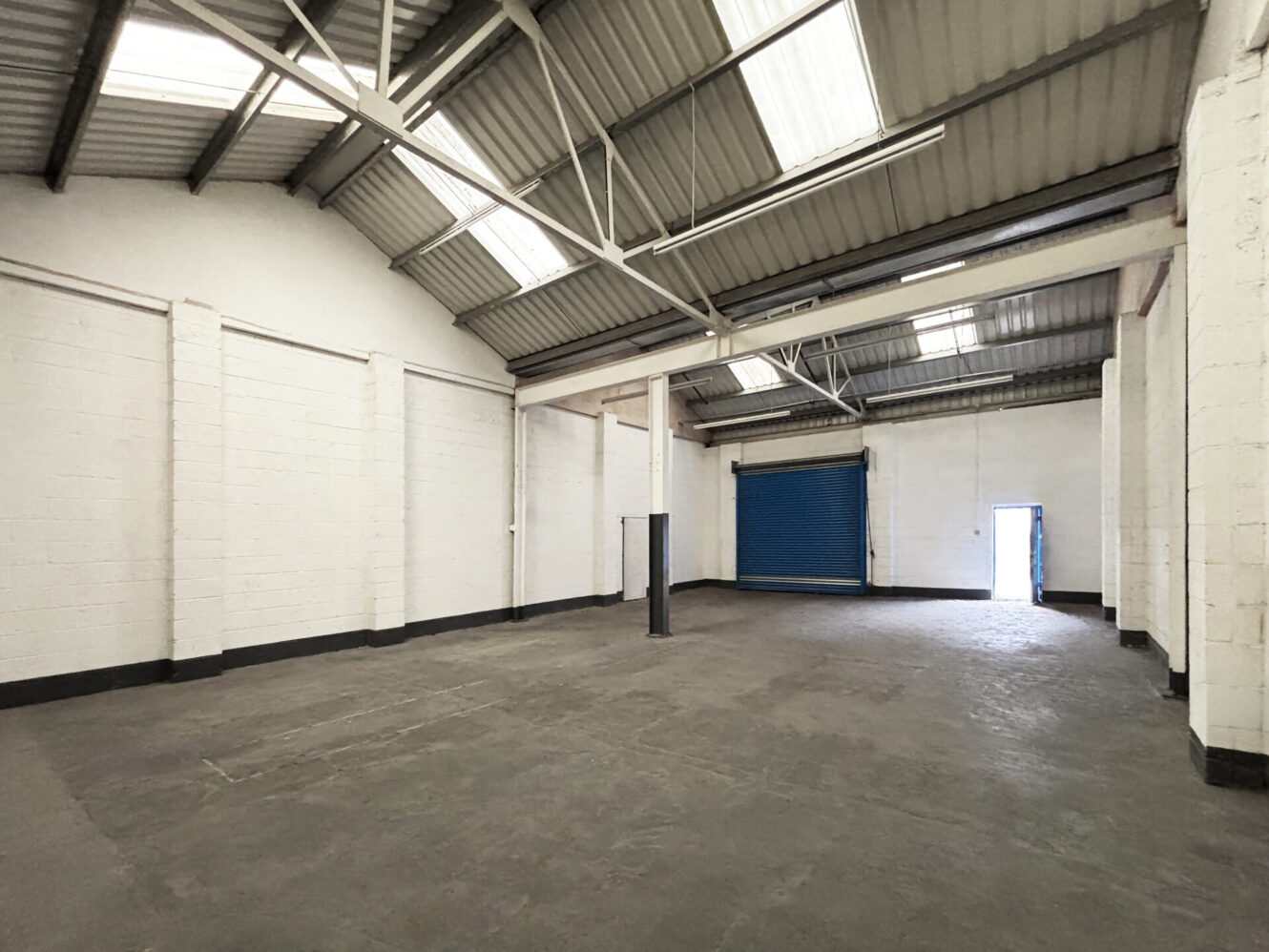 Interior of an empty industrial warehouse with a concrete floor, high ceiling, fluorescent lights, and a partially open blue roller shutter door.
