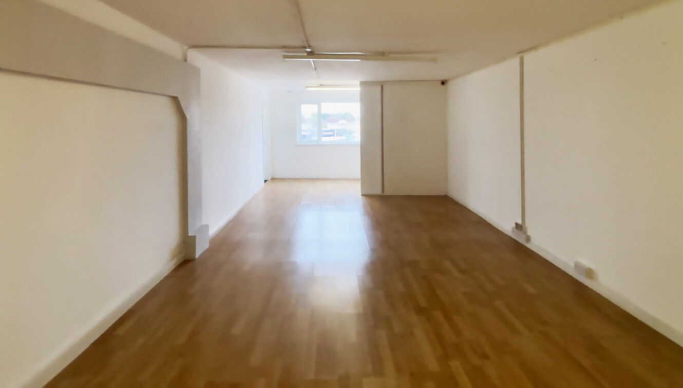 Empty rectangular room with white walls, wooden flooring, ceiling lights, and a window at the far end letting in natural light.
