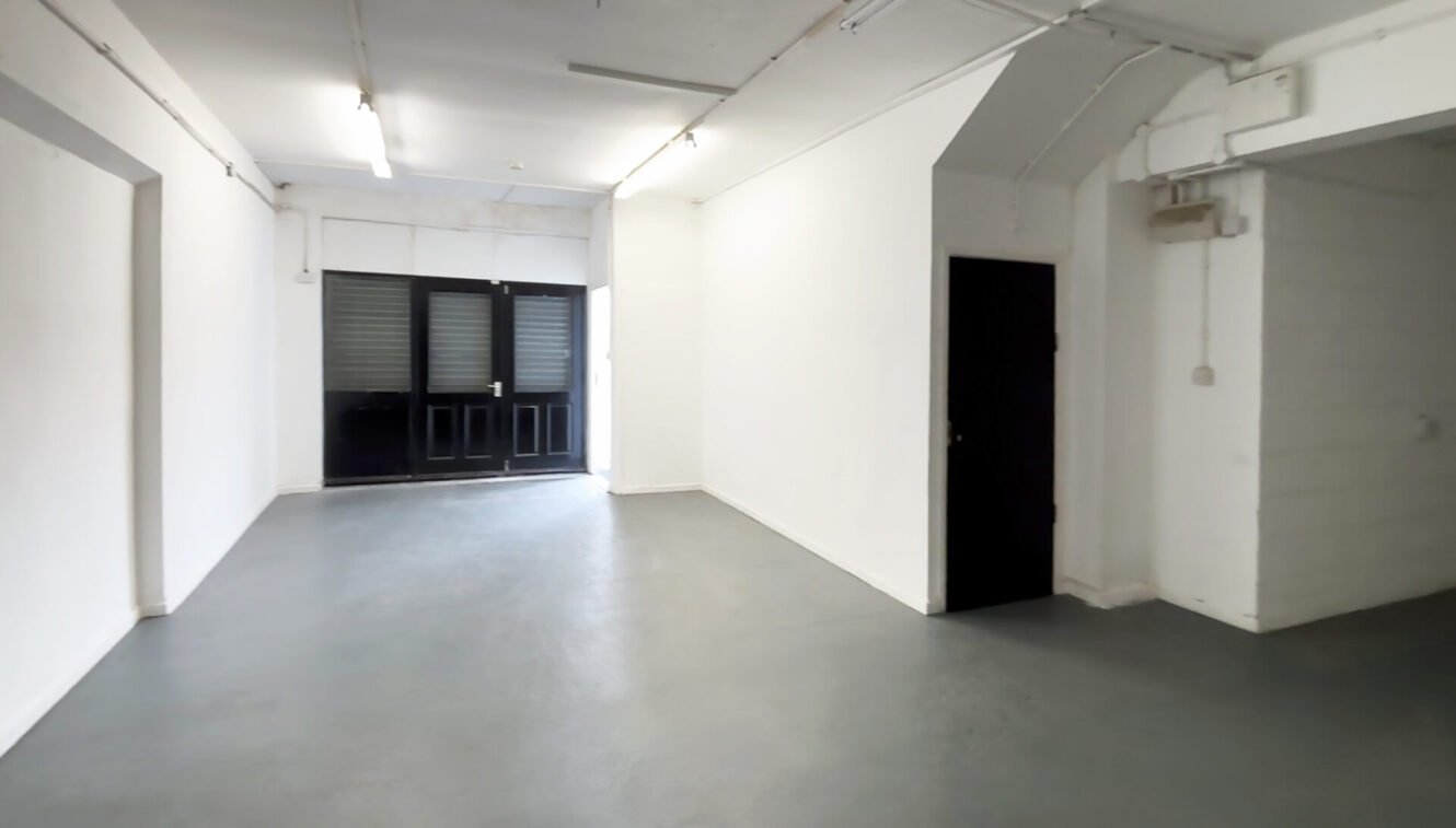 Empty room with white walls, grey floor, overhead fluorescent lights, a black door with windows at the far end, and a smaller black door on the right.