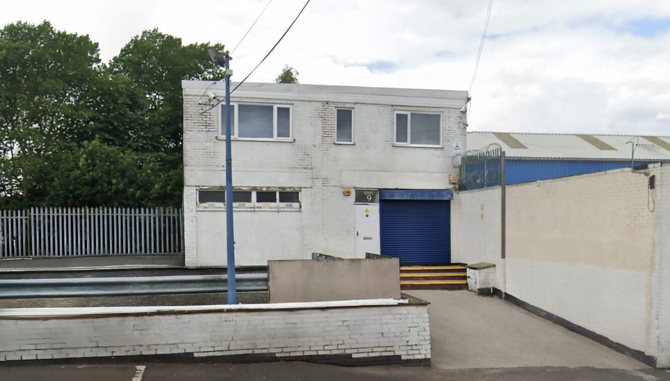 A white industrial building with a blue roller shutter door, loading bay, metal fence, and trees at the back under a partly cloudy sky.