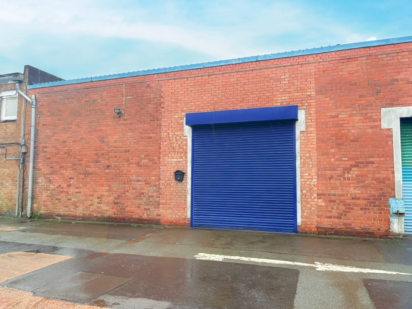 A red brick industrial building with a large closed blue roller shutter door, wet pavement, and cloudy sky overhead.