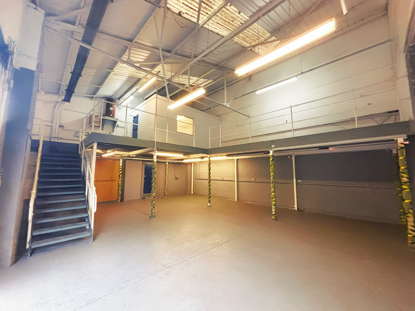 Industrial warehouse interior with mezzanine, metal staircase on the left, exposed beams, clear floor, and fluorescent lighting.