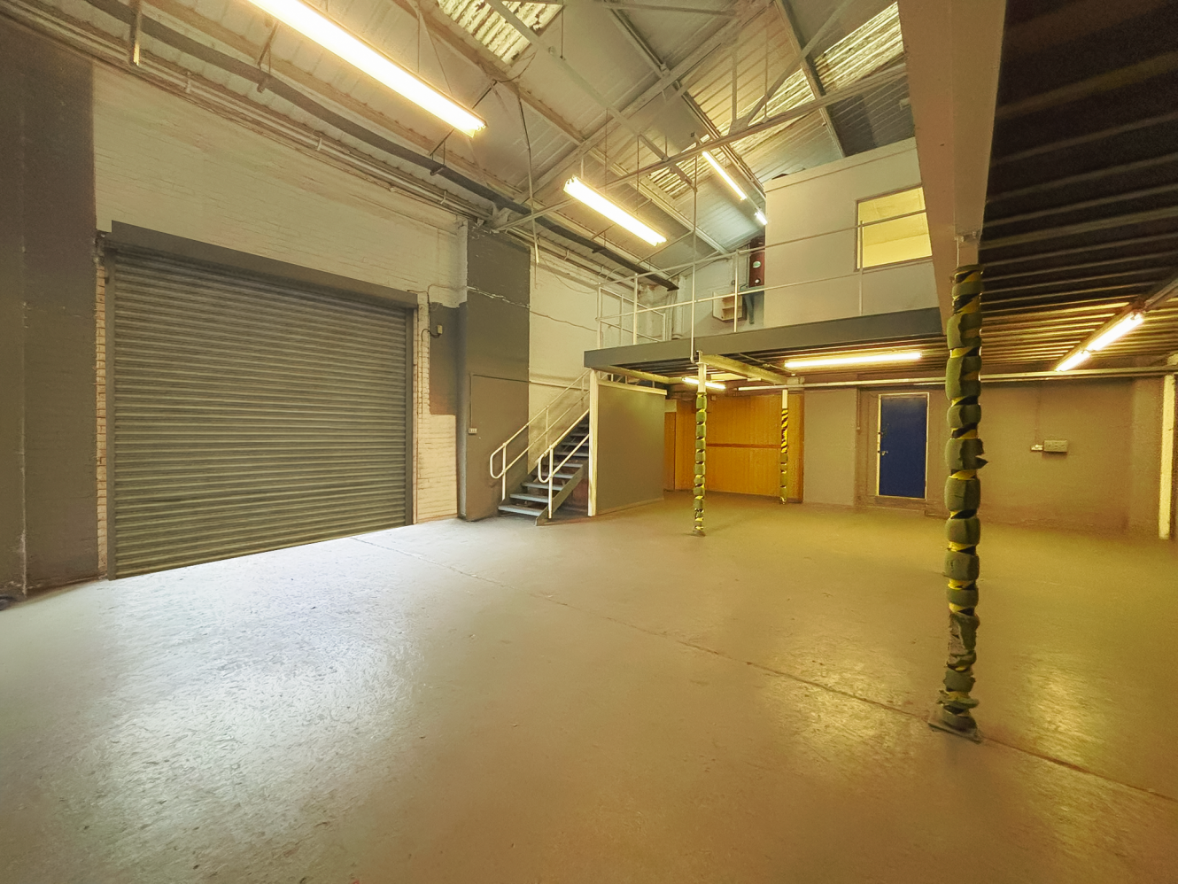 Spacious industrial warehouse interior with concrete floors, metal staircase, roller shutter door, mezzanine level, and fluorescent lights.