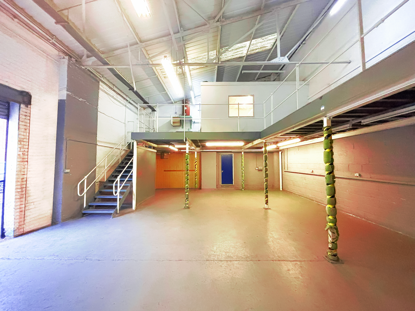Interior of an empty industrial warehouse unit with mezzanine level, metal stairs, exposed ceiling, and a blue door at the back.