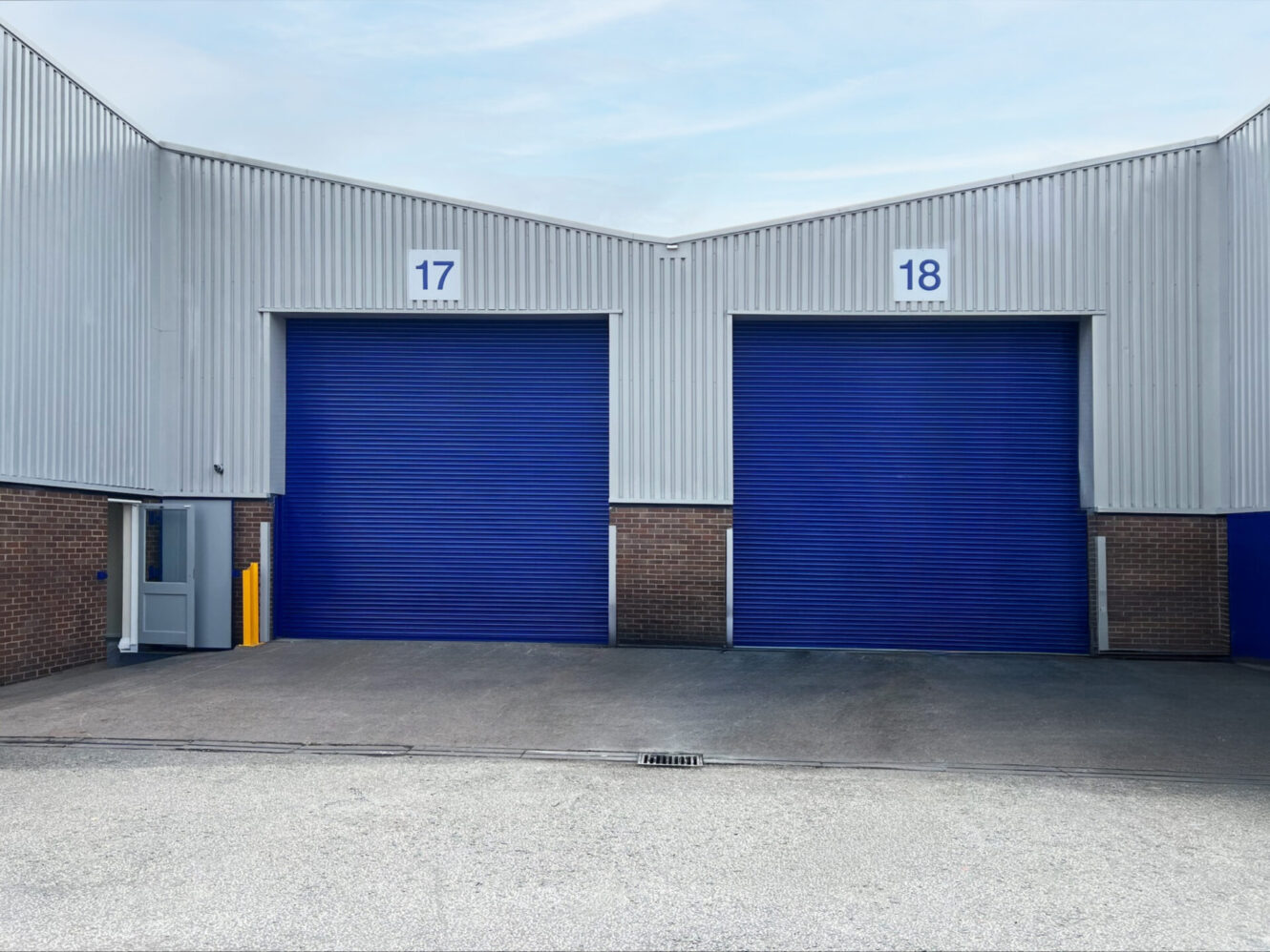 Two industrial warehouse units with blue roller shutters labelled 17 and 18, a small entry door on the left, and a paved area in front.