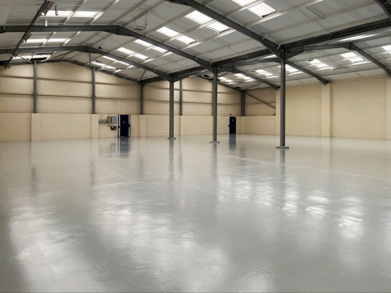 Large empty industrial warehouse with polished grey floor, beige walls, metal roof beams, fluorescent lights, and a few blue doors.