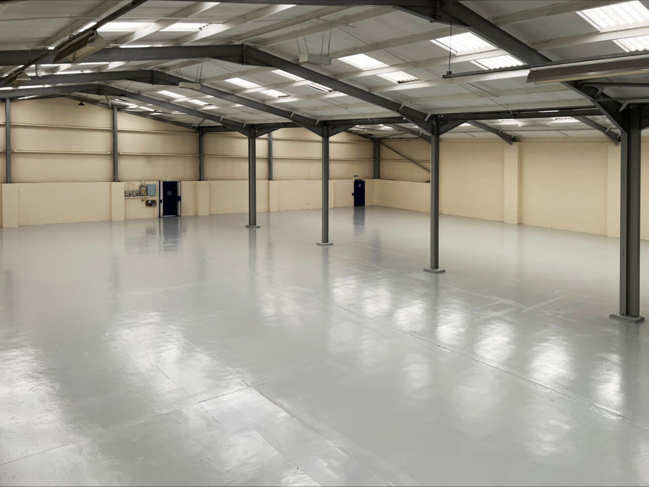 Interior of a spacious, empty industrial warehouse featuring polished concrete floors, steel beams, beige walls, and blue doors.