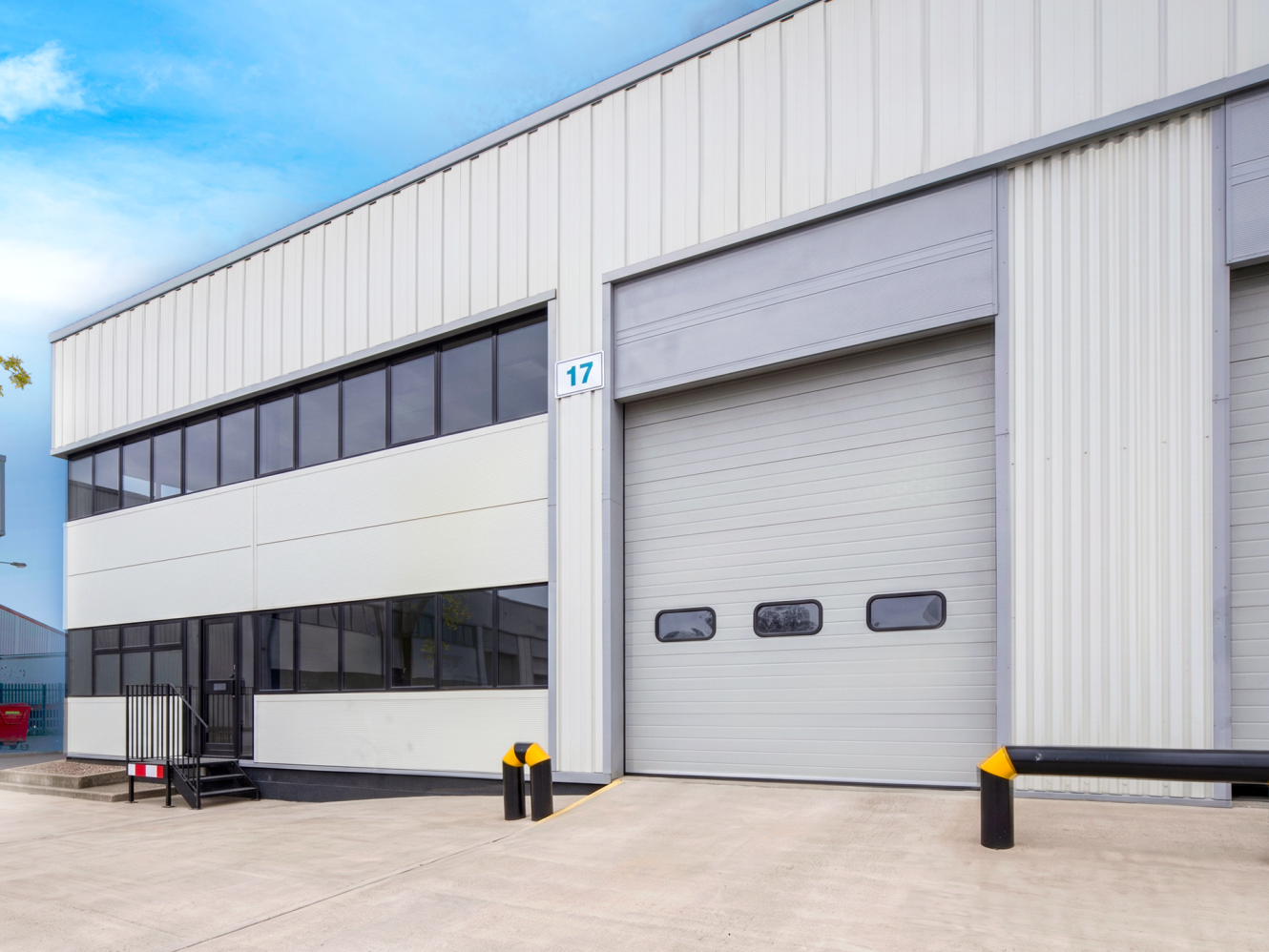 Exterior of a modern industrial warehouse with a large roller shutter door, windows above, and safety bollards in front; number 17.